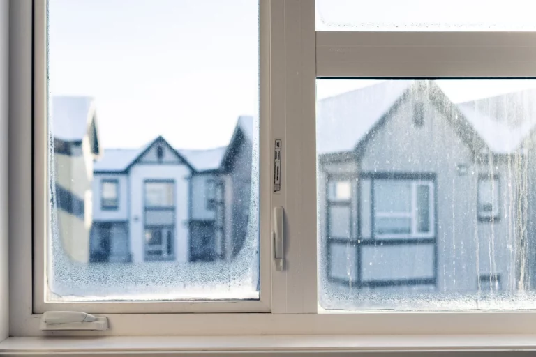A large window is frozen over on a frigid winter morning in Canada. The temperature outside is -30 degrees Celsius and the condensation has frozen over the glass panels. There is snow outside the window and the sun is slowly rising.